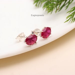 Puede incluir: Un par de pendientes de plata con piedras preciosas ovaladas rojas. Los pendientes están colocados sobre un fondo blanco.