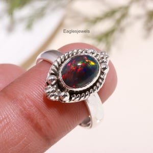 Op de afbeelding: Een zilveren ring met een zwarte opaal-edelsteen, gezet in een gedetailleerde zilveren setting. De opaal heeft een regenboog van kleuren, waaronder rood, groen en blauw.