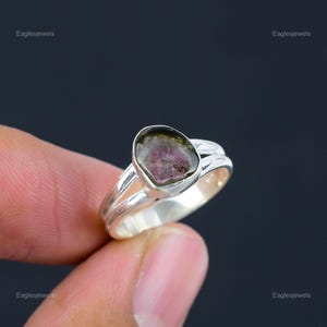 May include: A silver ring with a green, pink, and black gemstone. The gemstone is set in a silver band with a split band design.