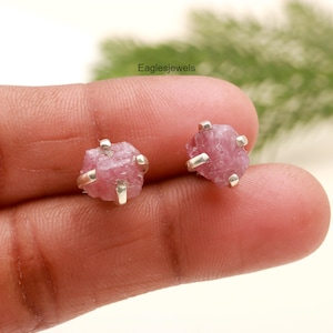 May include: A pair of pink tourmaline stud earrings. Each earring features a rough-cut, pink gemstone held in place by four silver prongs. The earrings are displayed on a person's finger. The text "eaglesjewels" is visible in the background.