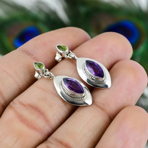 Peut inclure: Boucles d'oreilles pendantes en argent avec des pierres d'améthyste ovales et de petits accents de péridot. Les boucles d'oreilles ont une bordure texturée détaillée. L'arrière-plan est flou.
