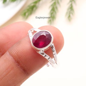 May include: A silver ring featuring a large, oval-cut ruby gemstone. The ring has a split shank design with geometric detailing. The ruby is a deep, rich red color, and the ring is held by a finger. The text "Eaglesjewels" is visible.