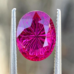 Puede incluir: Una piedra preciosa ovalada de color fucsia vibrante con un complejo patrón de facetas radiales en forma de estrella. La piedra está sujeta por unas pinzas de metal plateado, mostrando su color rosa intenso y sus cortes geométricos precisos.
