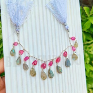 May include: A handmade necklace featuring pink gemstone beads and leaf-shaped pendants. The necklace is strung on a silver wire and has two white tassel accents. The leaf pendants have a mix of green, blue, and gray tones.