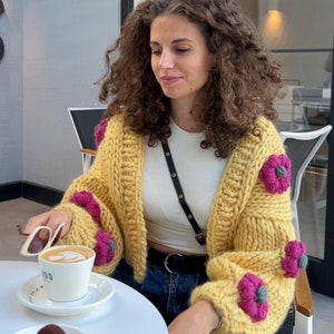 Puede incluir: Una mujer con un cárdigan amarillo mostaza grueso adornado con grandes flores rosas y verdes. Está sentada a una mesa blanca con una taza de café y un pequeño postre. El cárdigan es una pieza llamativa.