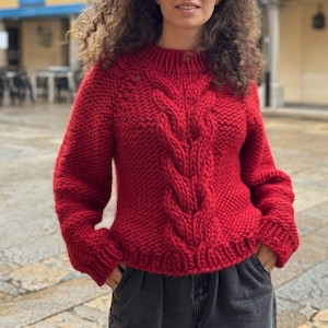 Könnte beinhalten: Ein leuchtend roter Strickpullover mit einem Zopfmuster in der Mitte. Der Pullover hat einen hohen Ausschnitt und lange Ärmel. Das dicke Strickwerk wirkt warm und gemütlich, ideal für kühleres Wetter. Der Pullover ist einfarbig.