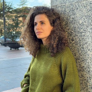 Peut inclure: Un pull vert tricoté avec un col montant et des manches longues. Le pull est de couleur unie et semble être fait d'un fil doux et texturé. La personne porte un jean bleu.