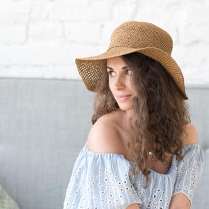 Op de afbeelding: Een vrouw draagt een bruine geweven zonnehoed met een brede rand en een lichtblauwe off-the-shoulder jurk. De hoed heeft een ronde kroon en een geperforeerd ontwerp op de rand. De jurk heeft witte eyelet details.