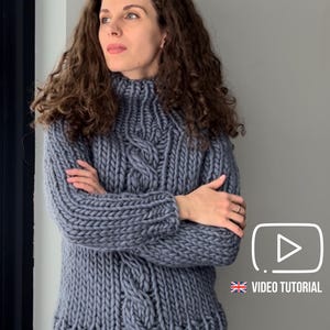 Puede incluir: Un suéter gris de punto grueso con cuello alto y detalles de punto trenzado. El suéter lo lleva una persona con pelo largo, rizado y castaño. La imagen incluye un icono de tutorial de vídeo en la esquina inferior derecha.