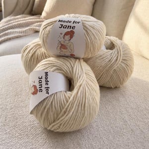 May include: Four ivory-colored yarn skeins are stacked on a neutral-toned surface. Each skein has a label that reads "Made for Jane" with an illustration of a person crocheting. The yarn appears soft and thick, ideal for knitting or crocheting projects.