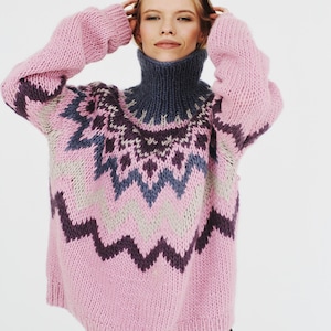 Könnte beinhalten: Ein rosa, übergroßer Strickpullover mit einem hohen, dunkelgrauen Rollkragen. Der Pullover hat ein geometrisches Muster in Lila-, Blau- und Grautönen auf der Brust und den oberen Ärmeln. Die Ärmel sind lang und der Pullover scheint aus einem groben Strick zu sein.