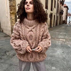 May include: A chunky knit, blush-colored sweater with a high, ribbed collar and intricate cable knit detailing. The sweater has a relaxed fit with long sleeves. The person is wearing gray pants and holding sunglasses.