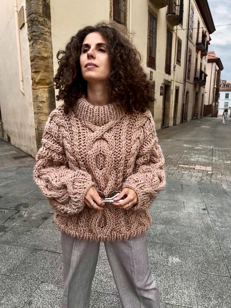 May include: A chunky knit, blush-colored sweater with a high, ribbed collar and intricate cable knit detailing. The sweater has a relaxed fit with long sleeves. The person is wearing gray pants and holding sunglasses.