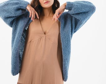 May include: A blue knitted cardigan with long sleeves is worn over a tan dress. The cardigan has a textured appearance and is open at the front. The dress has a V-neck and a tiered skirt. The background is white.