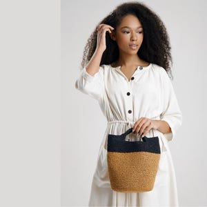 Op de afbeelding: Een crèmekleurige midi-jurk met een knoopsluiting aan de voorzijde en een getailleerde taille, gecombineerd met een tweekleurige handtas. De handtas heeft een zwart bovendeel en een beige onderkant, gemaakt van gevlochten materiaal met twee handvatten.