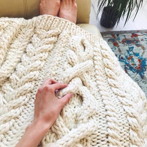 May include: Cream-colored, chunky knit blanket with a cable knit design. The blanket is draped over a person's legs and feet. The thick yarn creates a cozy, textured appearance, perfect for home decor and comfort.