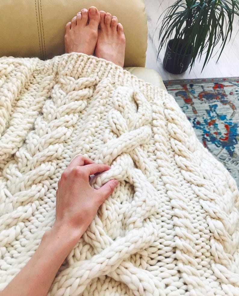 May include: Cream-colored, chunky knit blanket with a cable knit design. The blanket is draped over a person's legs and feet. The thick yarn creates a cozy, textured appearance, perfect for home decor and comfort.