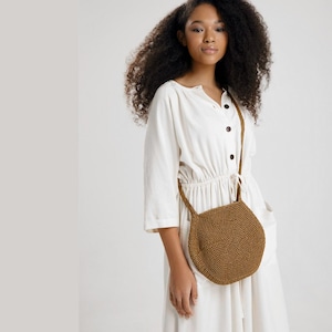 May include: A brown, crocheted crossbody bag with a round, geometric shape. The bag has a long strap and is worn over the shoulder. The bag is made of a textured, woven material.