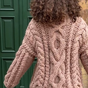 May include: A chunky knit sweater in a soft, muted rose color. The sweater features a prominent cable knit design on the front and sleeves, with a high neckline. The texture of the knit is clearly visible, suggesting a warm and cozy garment.