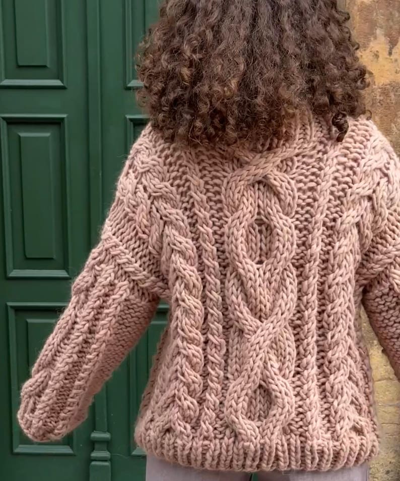 May include: A chunky knit sweater in a soft, muted rose color. The sweater features a prominent cable knit design on the front and sleeves, with a high neckline. The texture of the knit is clearly visible, suggesting a warm and cozy garment.