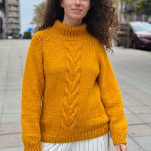 Könnte beinhalten: Senfgelber Zopfstrickpullover mit hohem Rippkragen. Der Pullover hat ein zentrales Zopfmuster und lange Ärmel. Das Model trägt den Pullover im Freien. Der Pullover ist aus einem dicken, warmen Material gefertigt.