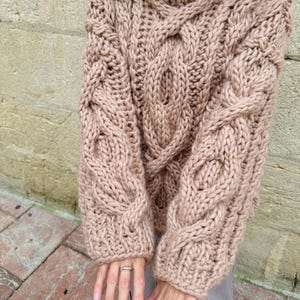May include: A chunky knit sweater in a light brown color. The sweater features a cable knit design on the sleeves and front, with a high, ribbed turtleneck. The sleeves are long and the overall style is oversized and cozy.
