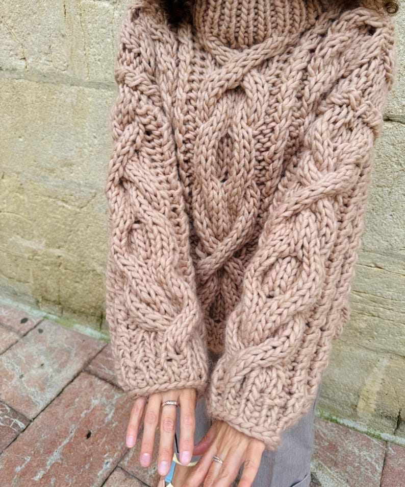 May include: A chunky knit sweater in a light brown color. The sweater features a cable knit design on the sleeves and front, with a high, ribbed turtleneck. The sleeves are long and the overall style is oversized and cozy.