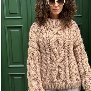 May include: A chunky, beige cable-knit sweater with a high neckline. The sweater features intricate cable patterns on the front and sleeves. The sleeves are long and the fit is oversized. The background is a green door.