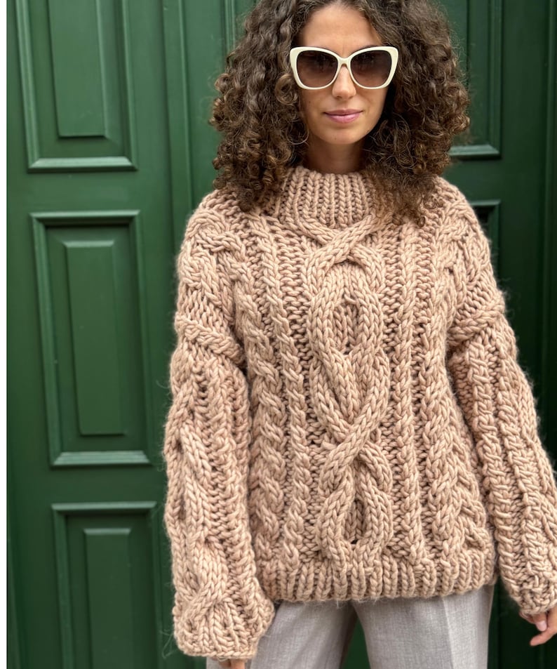 May include: A chunky, beige cable-knit sweater with a high neckline. The sweater features intricate cable patterns on the front and sleeves. The sleeves are long and the fit is oversized. The background is a green door.