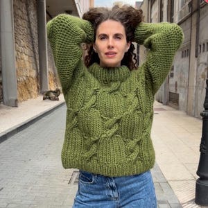 Könnte beinhalten: Ein grob gestrickter olivgrüner Pullover mit einem markanten Zopfmuster, langen Ärmeln und einem hohen Stehkragen. Das Kleidungsstück ist aus dickem Garn gefertigt und wird mit einer blauen Jeans kombiniert, was eine strukturierte Strickoptik zeigt.