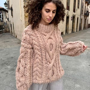 May include: A chunky knit sweater in a soft, muted rose color. The sweater features a high, ribbed collar and intricate cable knit detailing down the front and sleeves. The sleeves are long and have a slightly puffed shape. The sweater is made of a thick, warm yarn.