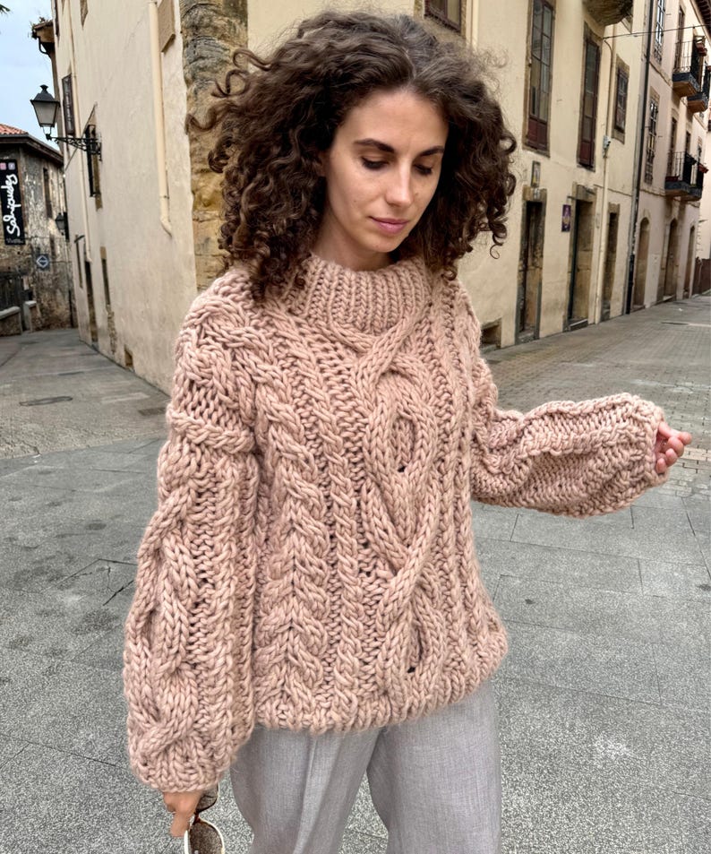 May include: A chunky knit sweater in a soft, muted rose color. The sweater features a high, ribbed collar and intricate cable knit detailing down the front and sleeves. The sleeves are long and have a slightly puffed shape. The sweater is made of a thick, warm yarn.