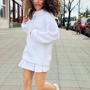 The photo shows a girl in a knitted white sweater. She is standing on a city street.