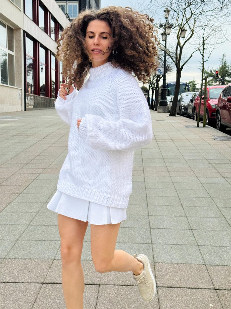 The photo shows a girl in a knitted white sweater. She is standing on a city street.