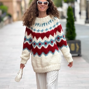 Könnte beinhalten: Ein cremefarbener, übergroßer Pullover mit einem geometrischen Muster in Rot und Hellblau. Der Pullover hat einen gerippten Ausschnitt und Bündchen. Die Person trägt eine weiße Sonnenbrille und eine gestreifte Hose und hält eine weiße Handtasche.