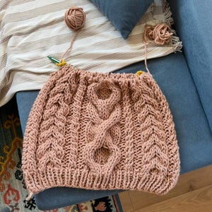 May include: A hand-knitted, blush-colored garment with a prominent cable-knit design. The piece is in progress, with yarn balls and needles visible. The texture of the knitted fabric is clearly defined, showcasing the craftsmanship. The item is likely a sweater or a similar article of clothing.