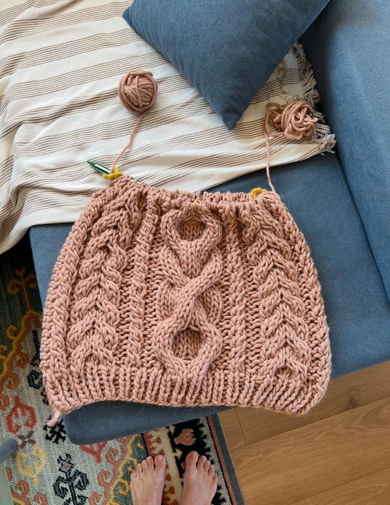 May include: A hand-knitted, blush-colored garment with a prominent cable-knit design. The piece is in progress, with yarn balls and needles visible. The texture of the knitted fabric is clearly defined, showcasing the craftsmanship. The item is likely a sweater or a similar article of clothing.