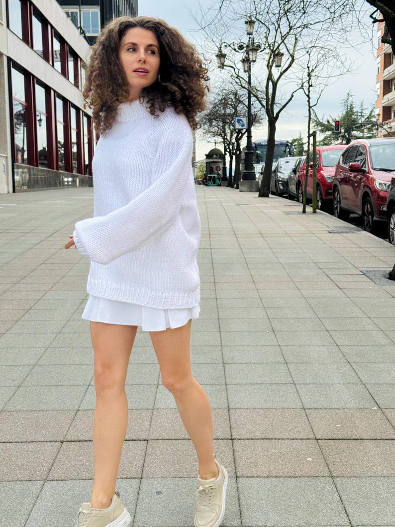 The photo shows a girl in a knitted white sweater. She is standing on a city street.