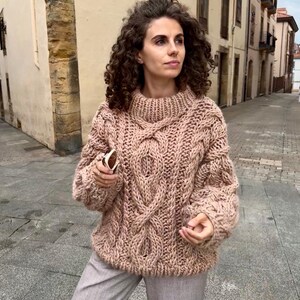 May include: A chunky knit sweater in a soft, muted rose color. The sweater features a high, ribbed collar and intricate cable knit detailing across the chest and sleeves. The oversized design suggests a cozy, warm garment.
