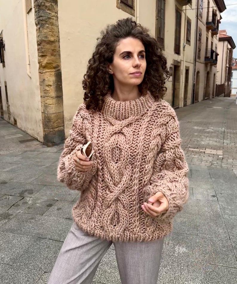 May include: A chunky knit sweater in a soft, muted rose color. The sweater features a high, ribbed collar and intricate cable knit detailing across the chest and sleeves. The oversized design suggests a cozy, warm garment.