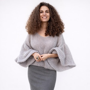Könnte beinhalten: Eine Frau trägt einen hellgrauen, übergroßen Strickpullover mit weiten Glockenärmeln und einem V-Ausschnitt. Sie trägt auch einen grauen Bleistiftrock. Der Pullover hat ein weiches, strukturiertes Aussehen. Die Frau lächelt und trägt Ringe.