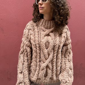 May include: A chunky, beige cable-knit sweater with a high neckline. The sweater features thick, textured yarn and a prominent cable design running down the front and sleeves. The sleeves are long and the overall style is oversized.