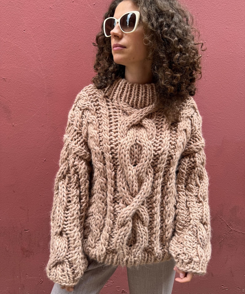 May include: A chunky, beige cable-knit sweater with a high neckline. The sweater features thick, textured yarn and a prominent cable design running down the front and sleeves. The sleeves are long and the overall style is oversized.