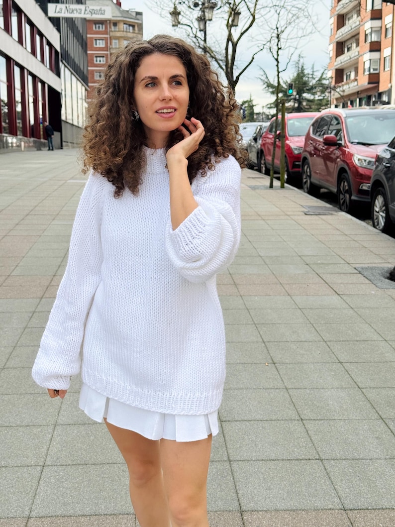 The photo shows a girl in a knitted white sweater. She is standing on a city street.
