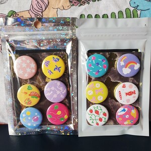 Op de afbeelding: Twee sets ronde, kleurrijke pinback-knoppen met verschillende ontwerpen. De eerste set bevat een witte ster, een gele vlinder, een paarse en roze swirl, een gele taco, een blauwe planeet en een roze en groene swirl. De tweede set bevat een blauwe lolly, een paarse regenboog, een gele strik, een rode snoepstok, een rode aardbei en een roze en blauwe fee.