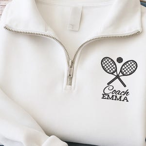 Puede incluir: Sudadera blanca de entrenador de tenis con cremallera plateada y el texto "Coach EMMA" debajo de un gráfico de raquetas de tenis cruzadas y una pelota. La sudadera está doblada sobre unos vaqueros azules.
