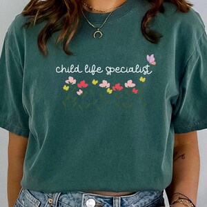 May include: A teal blue t-shirt with the text "child life specialist" in white script. The shirt has a floral design with pink, yellow, and white flowers.