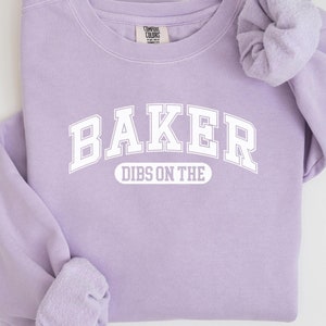 May include: A lavender sweatshirt with the text "BAKER DIBS ON THE" in white. The sweatshirt has a crew neck and long sleeves.