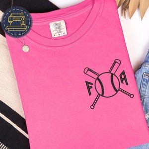 Op de afbeelding: Een felroze T-shirt met korte mouwen en een zwart geborduurd ontwerp op de linkerborst. Het ontwerp bestaat uit twee gekruiste honkbalknuppels achter een honkbal, met de letters F en A aan weerszijden van de bal geplaatst.