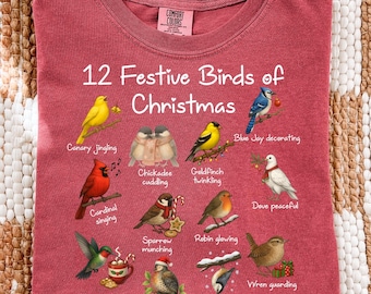 May include: A maroon Comfort Colors t-shirt featuring the text "12 Festive Birds of Christmas" with illustrations of various birds. The shirt has a crew neck and short sleeves. The birds are depicted in a festive holiday theme.